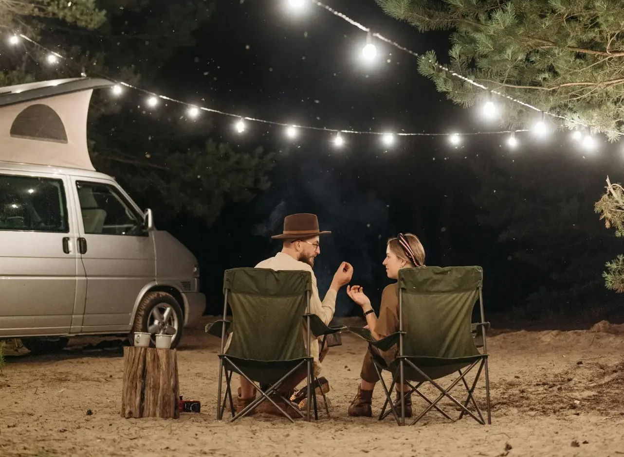 A couple enjoys a quiet night camping under string lights by a van amidst green trees.